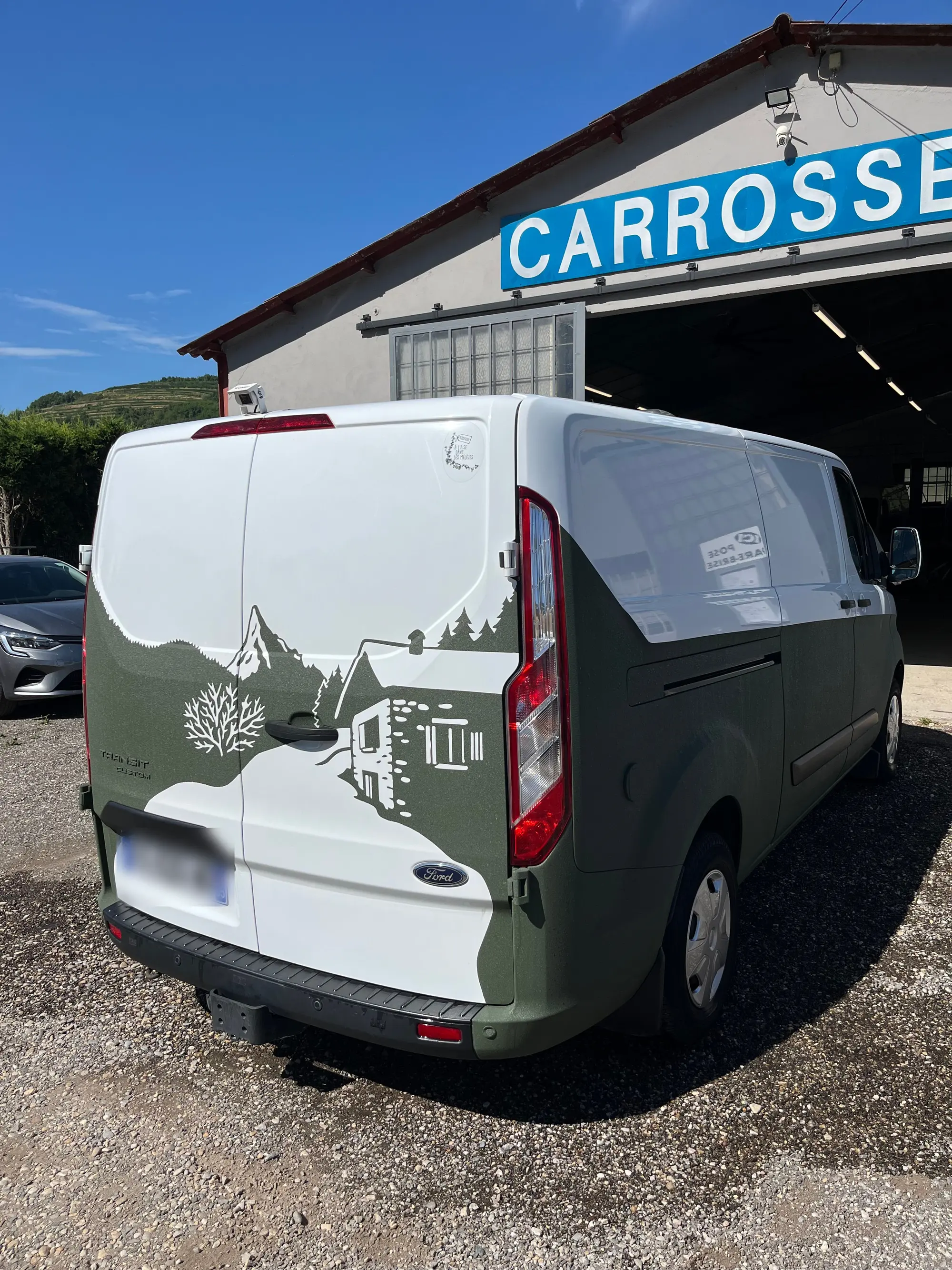Fourgon Ford blanc et vert kaki décoré d’un paysage de montagne, devant l’enseigne de l’atelier